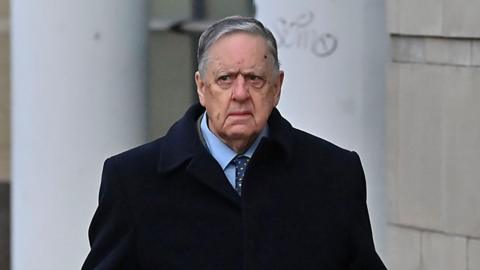 William Lloyd-Lavery walking outside. He is wearing a navy jacket over a light blue shirt with a navy tie with multi-coloured spots on it. He has grey hair. 