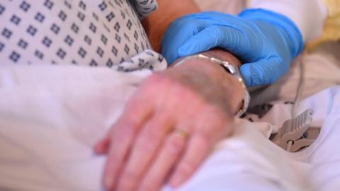 A hand wearing a blue latex glove is on a female hand in a comforting manner. The woman is in a bed.