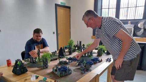 Two gamers playing Warhammer 40k. One man is standing up and is moving a small model of a soldier across a table littered with scenery. A second man is sitting down looking a the battlefield.