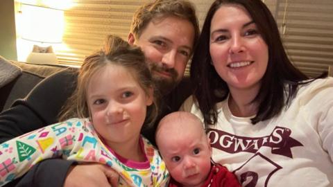 The photo shows Tom with his wife and children on a sofa. Tom has short brown hair and a well trimmed beard. He is wearing a black jumper. His wife has shoulder length dark brown hair. She is wearing a white jumper.