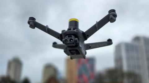 A drone in the air. The grey four-pronged equipment is in front of the blurred outline of tower blocks