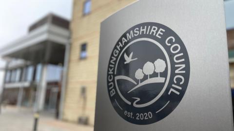 A view of a grey sign with the Buckinghamshire Council logo on it. A building can be seen behind it.