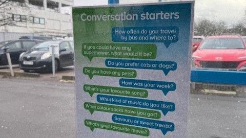 A list of conversation starters displayed on the window of a chatty bus. There are questions such as "How often do you travel by bus and where to?" listed on it. There are cars seen parked through the window.