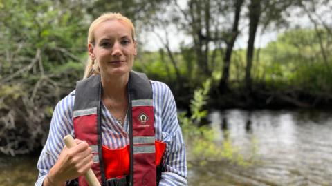 Alice smiles at the camera. She has her blonde hair tied up and wears orange river waders, a blue and red life jacket, and a blue striped shirt underneath. She holds a wooden pole and stands in front of a river, blurred in the background.
