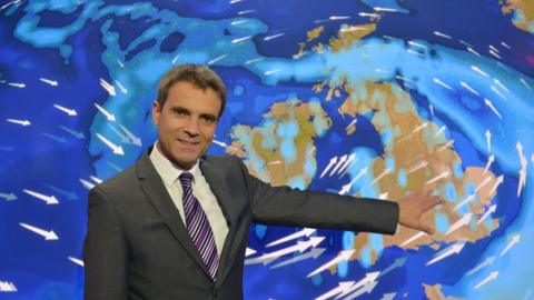 Weather presenter in a suit pointing towards weather map of UK.