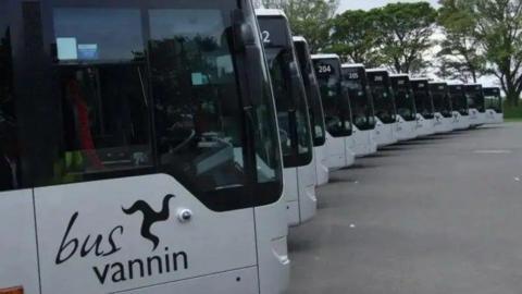 The front of a row of buses stretching round in a semi-circle, They are silver with dark tinted windows and have Bus Vannin alongside a triskelion on the side.