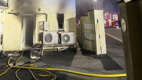 The side entrance of a building with a doorway blackened by smoke. The outside of the building has large fan units and an electricity box which is open. A yellow fire hose is snaked on the ground in front of it leading in through the doorway, and a lit up fire engine can be see on the road in the background.