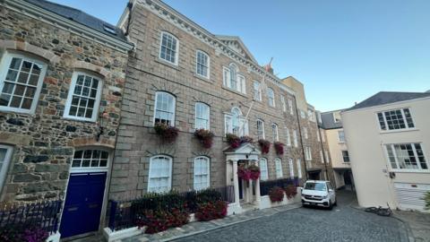 A three storey granite government building, with rows of arched windows. Handing baskets of flowers sit beneath many of them.