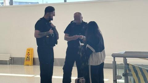 Two men in dark uniforms from Home Office Enforcement question a woman at an airport. Although both men's faces are blurred, one is tall with dark hair and a beard while the other is balding. The woman, who has long black hair, has her back turned to the camera. 
