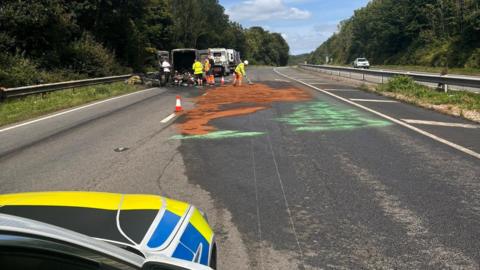 Van fire on A380, with brown and green colours on carriageway and workers in the background near burnt-out vehicle.