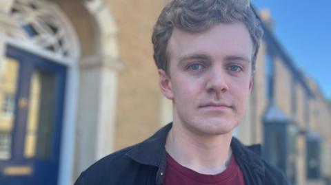 Sam Carling looking down the lens of a camera. He is dressed casually. Behind him is a line of terraced houses.