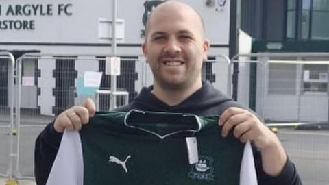 A person holds up a Plymouth Argyle shirt outside Home Park stadium, with the Plymouth Argyle FC Superstore visible in the background. 