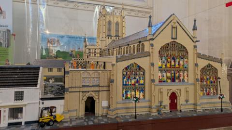 The outside of a model Lego replica of St Edmundsbury Cathedral with a spire and tower and stained glass windows.