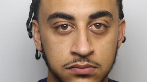 Custody photograph of a person with dark braided hair, shown from the shoulders up against a plain light background.