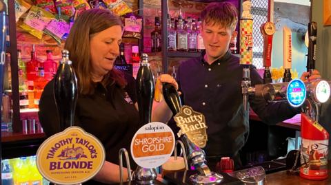 Julia Buckley has brown hair and is behind a bar pulling a pint of Butty Bach. A man is standing next to her. He has brown hair and is wearing a black shirt. Around them on the bar are various beer taps, with crisps on the wall behind them and shelves of spirits.