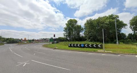 Google street view image of a three-laned roundabout, which has trees and grass in the middle.