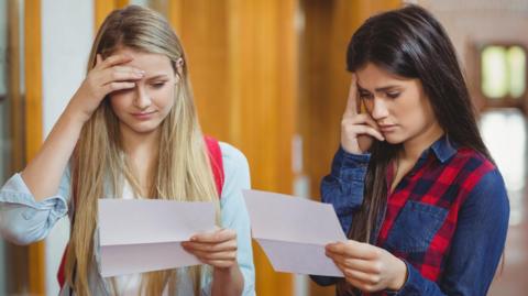 Two girls stand in a school corridor looking anxiously at sheets of paper they are holding. One hard blonde hair and the other had dark hair. 