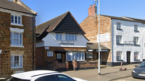 The Oak House Hotel which stretches across three buildings. A white and black car are also parked on the street and can be seen in the picture.