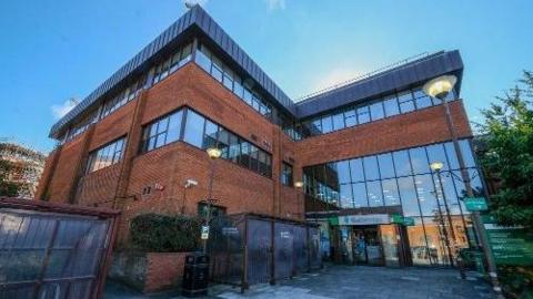 The West Berkshire Council building. It is a red brick building with a glass frontage and a flat foor. The building has four storeys. There are green signs above the entrance saying West Berkshire Council.