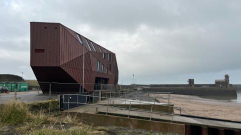 A general view of The Edge. The outer shell of the building appears complete and is surrounded by temporary fencing. It sits in front of the Golden Sands beach with the harbour wall visible in the distance. The building is an irregular geometrical shape covered in dark brown cladding.