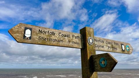 A Norfolk Coast Path fingerpost brightly lit in front of a partially cloud sky