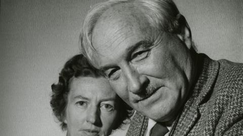 A black and white photo of Louis with his second wife Mary Leakey. He is close to the camera and has a small moustache, light coloured hair and is smiling with his mouth closed. His wife has short dark and curly hair. She further behind Louis and also looking at the camera. 