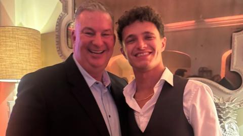 A white man with grey hair smiles laughing at the camera, next to a young gentlemen with brown hair and smile, white shirt and waist coat.