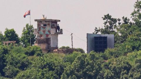 A blurry photo of a soldier standing on a tower, next to a giant loudspeaker. A North Korean flag is mounted next to the tower.
