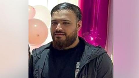 Naweed Hussain, standing in front of some balloons, wearing a dark top and coat, he has a dark beard and short dark hair. 