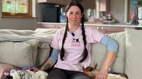 A woman has long brown hair tied in plaits and is wearing a pink t-shirt that says 'Heidi's Courageous Cat Foundation'. She is sitting on a beige sofa with a dog and cat by her side.