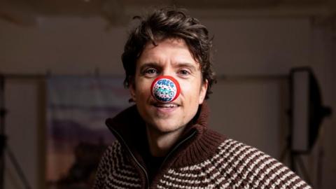 A close-up photo of Radio 1 DJ Greg James, he is wearing a brown and white stripped knitted jacket and has long curly brunette hair. He is staring directly at the camera and has a red nose on his nose that has a picture of a tandem bike on it and says Greg James.