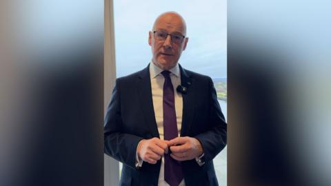 Scottish National Party leader John Swinney smiles at the camera as he stands in front of a wide window. He is wearing glasses, a maroon tie, white shirt and navy blue suit.