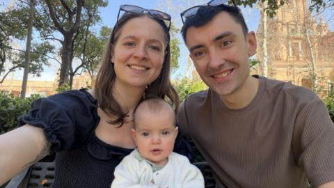 A woman with a black shirt is sitting next to a man in a brown jumper. There is an infant sitting in between the couple.
