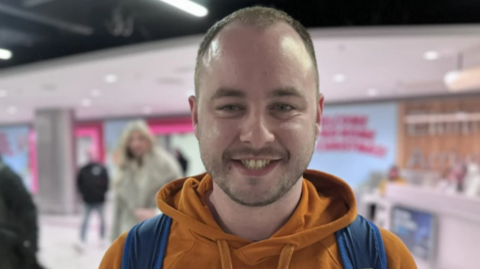 Craig Ballentine at Dublin Airport, he has short light hair and a beard. He is smiling at the camera and wearing an orange hoodie and a blue back pack 