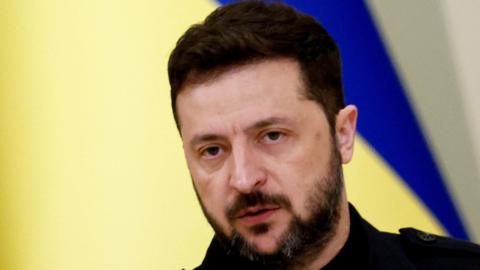 Zelensky - who has short, dark hair and a beard, dressed in a dark shirt, posed against a bold backdrop—a split field of yellow and blue, unmistakably mirroring the Ukrainian flag