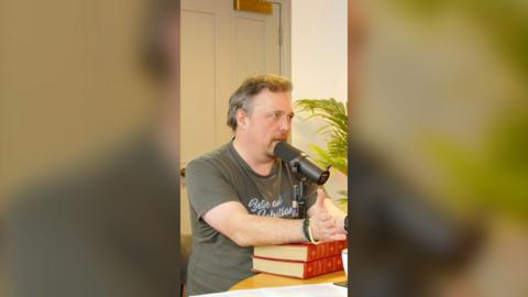 A man is sat at a table with a black mic in front of him that's resting on two red books.