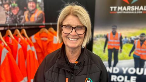 Jane Corlett smiling. She is standing next to orange high visibility jackets and A sign encouraging marshals to sign up.