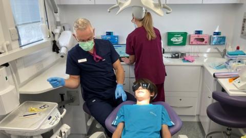 A 12-year-old boy sits in a dentist's chair. He wears protective goggles. The dentist wears scrubs and a mask and rubber gloves. In the background, an assistant prepares the equipment.