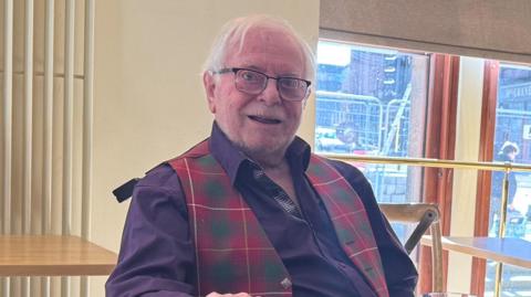Bob Mcfie sits indoors at a café or restaurant table, wearing a dark shirt with a tartan-style vest.