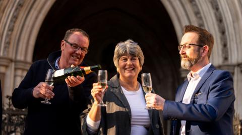 A woman stands in the middle of two men. She is wearing a dark jacket and white tee-shirt, one of the men wears a blue suit, the other is wearing a dark jumper. They are pouring a bottle of champagne. They are standing in front of an ornate, carved stone arch. 