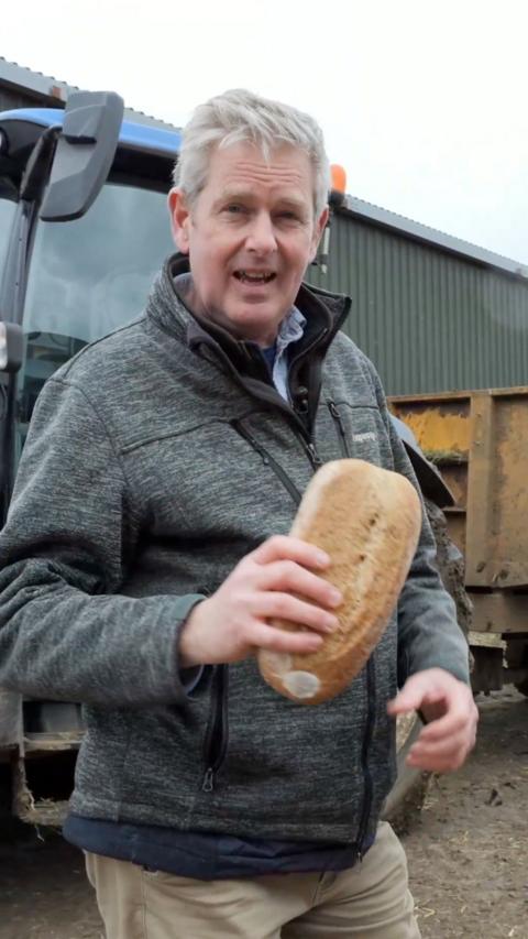 A man holding a baguette whilst standing in front of a tractor on a farm.