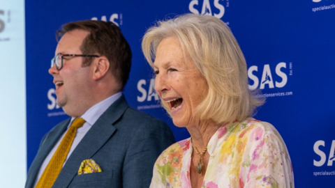 Bunny Campione laughing as she stands at the rostrum of the auction house, next to Thomas Forrester, who is wearing a suit and also laughing.