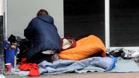 A rough sleeper wearing a blue jacket kneeling in a shop doorway with their back to the camera with an orange sleeping back and blankets and carrier bags  