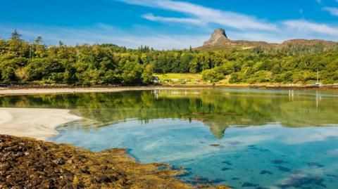 A scene on Eigg with a beach, and mirror-like calm sea. On the opposite shore is an area of trees and its landmark hill, The Sgùrr.