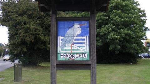 A village sign saying Herongate, with trees behind it and grass in front. The sign shows a heron with a blue sky. The sign is in a large wooden frame.