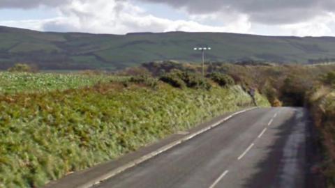 Glenfaba Road, which is a country road that runs alongside agricultural fields. Hills can be seen in the distance as the road dips out of view.