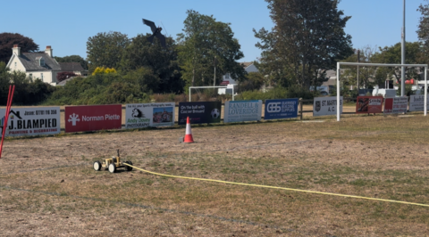 Grass field with goal on the right and a hose, a orange cone and a black crow scarecrow just in shot against the trees.