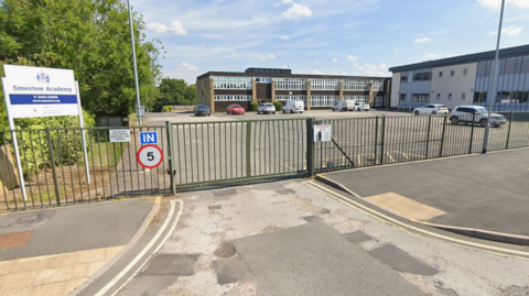 The metal school gates are closed with school buildings visible in the distance after a car park where some cars are parked. A sign bearing the school's name can be seen just inside the metal gates on the left of the entrance.
