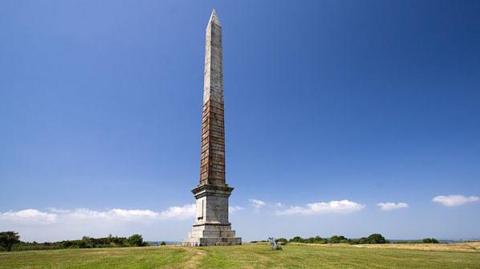 A wide shot of the Gilbert Memorial in front of a blue sky. The monument is very tall and weathered.