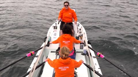 Three women in orange jumpers are rowing in a small white rowing boat on a grey sea with small waves.
